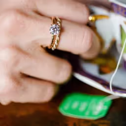 Close-up of a hand wearing a gold ring with a prominent round diamond, holding a tea bag with a green tag.