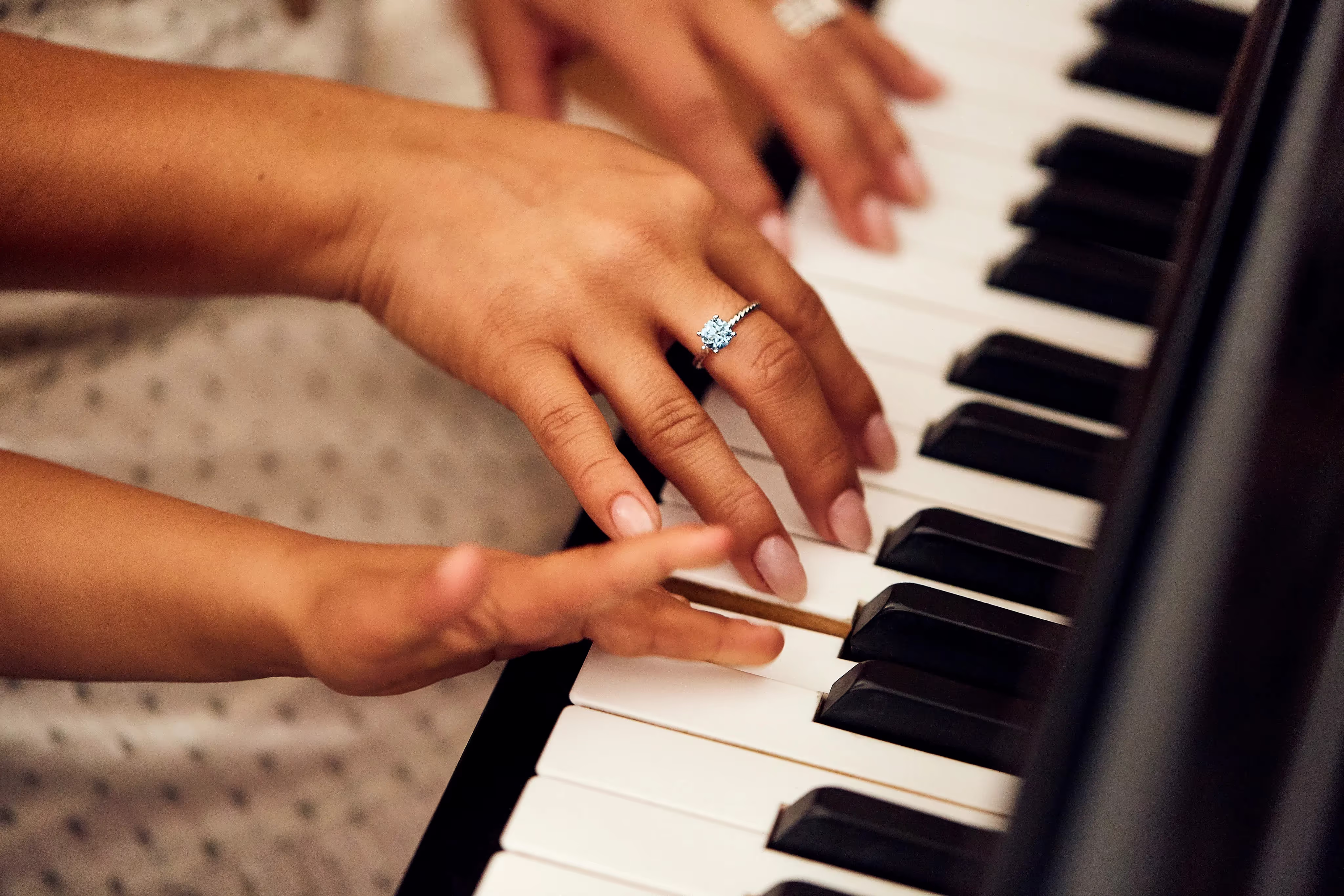 Close-up of hands playing piano keys, one hand wearing a sparkling engagement ring with a blue gemstone.