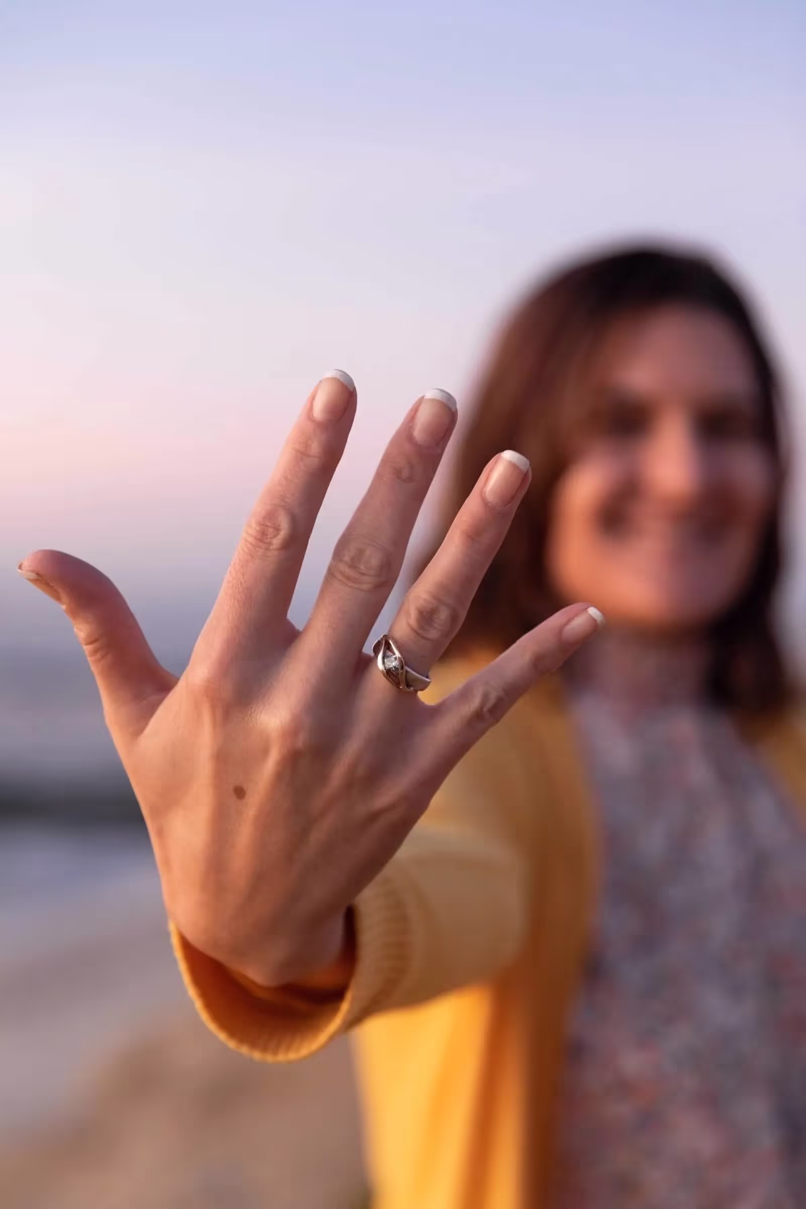 Close-up of a woman's hand with a diamond ring on her ring finger, blurred smiling woman in a yellow sweater in the background.