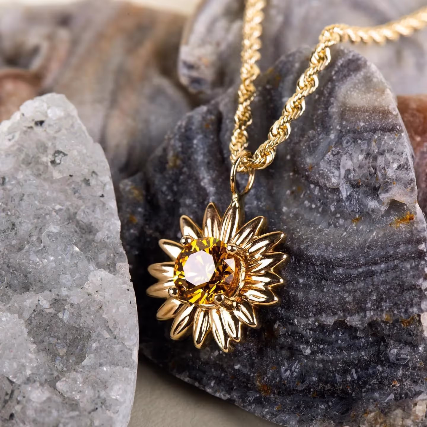 Gold necklace with a sunflower pendant featuring a yellow gemstone center, placed on textured gray and white rocks.