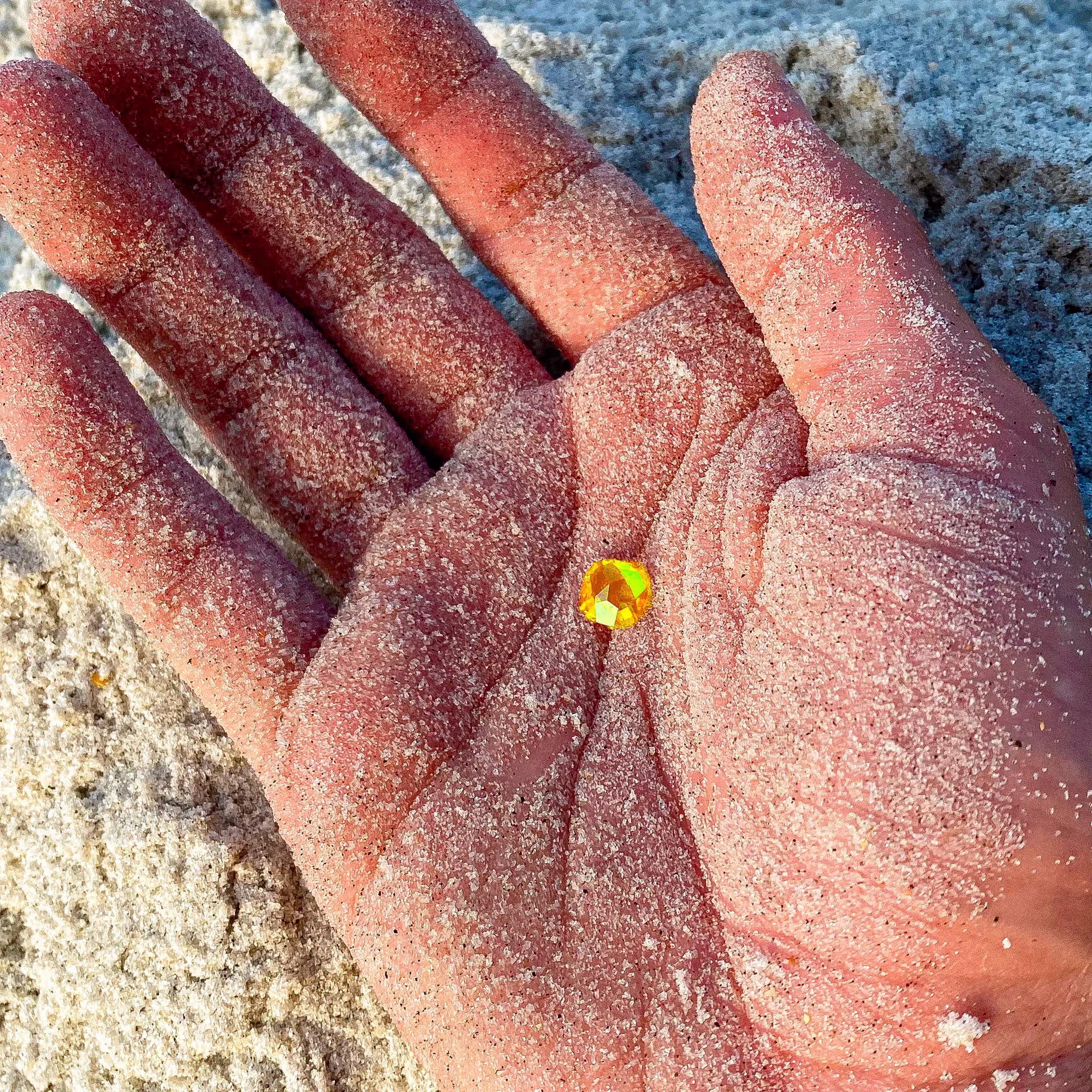 Close-up of a sand-covered hand holding a small yellow gemstone on a sandy surface.