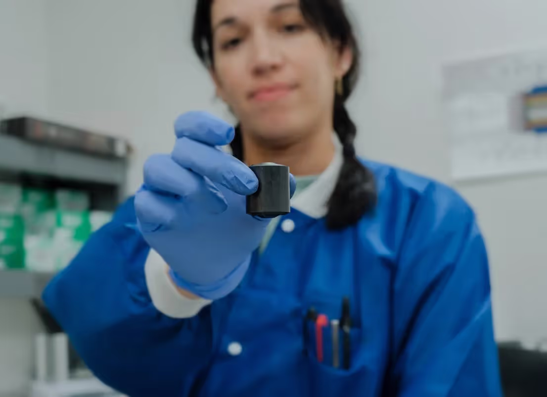 A scientist in a blue lab coat and gloves holding a small black container towards the camera.