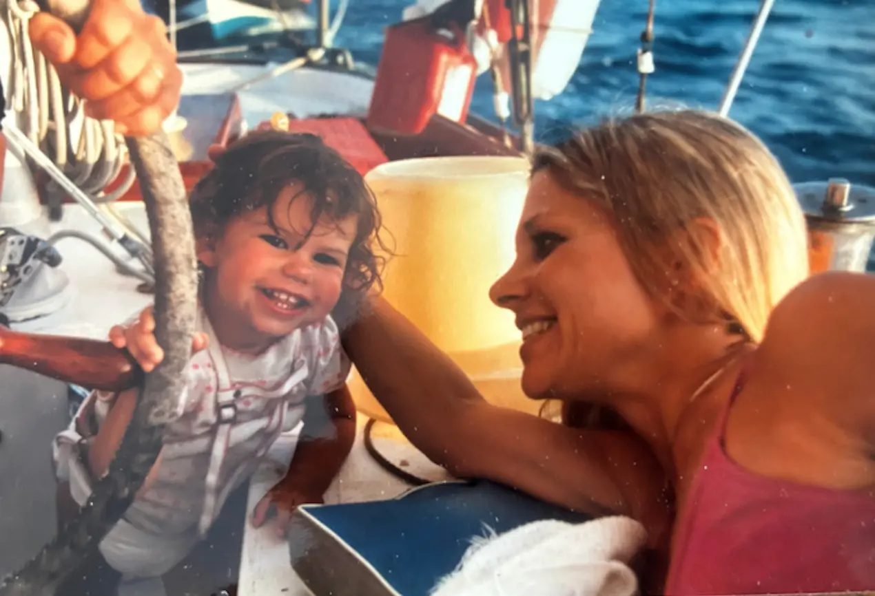 Vintage family photo of a mother and a smiling toddler at the tiller of a sailboat.
